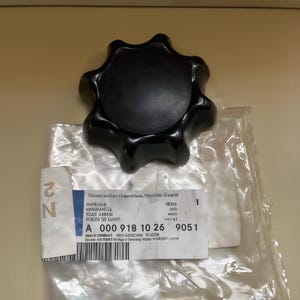 Può includere: Un pomello nero a forma di stella, probabilmente un componente per auto, è appoggiato su un'etichetta bianca e un imballaggio di plastica trasparente. L'etichetta presenta testo e numeri, tra cui "A 000 918 10 26". Il pomello è il soggetto principale, con una superficie liscia e lucida.