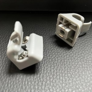 May include: Two light grey plastic car sun visor clips. One clip is open, and the other is closed. The clips have a rectangular shape with a curved hook and metal components. The background is a textured, dark grey surface.