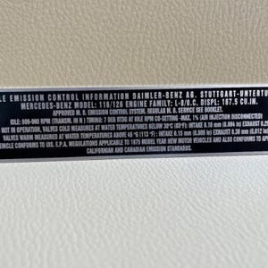 May include: A black and silver Mercedes-Benz vehicle emission control information plate. The plate includes technical specifications, model information, and regulatory compliance details. The text is in white, with the Mercedes-Benz logo on each end.
