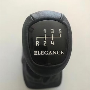 May include: Black gear shift knob with a leather-like texture. The top features a black panel with white markings for the gears: R, 1, 2, 3, 4, and 5. The word "ELEGANCE" is printed below the gear pattern.