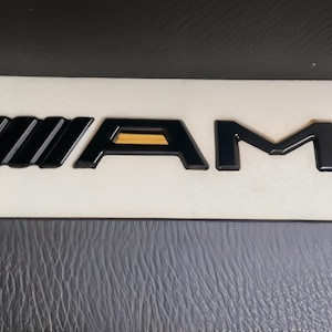 Op de afbeelding: Een zwart en goudkleurig AMG-embleem met een gestileerd schuin balkontwerp gevolgd door de letters A, M en G. De letters hebben een zwart frame met goudkleurige inzetstukken. Het embleem is gemonteerd op een rechthoekige witte basisplaat.