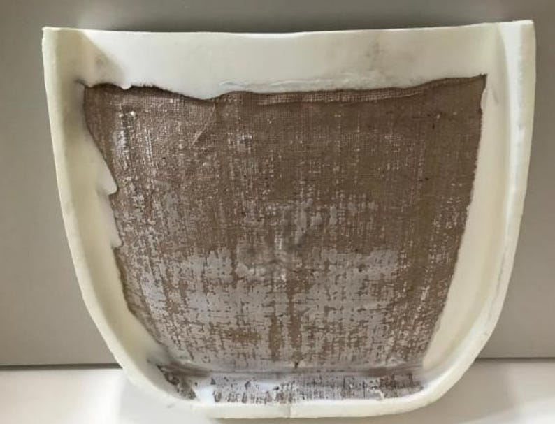 a white object with a textured surface, possibly a piece of furniture or a decorative item. The object appears to be made of a material that resembles burlap or burlap-like fabric, with a white base or frame. The object is placed against a plain white background, which highlights its details and textures.