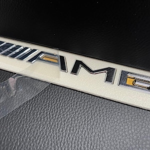 May include: Chrome AMG emblem on a white rectangular base, partially covered by a clear protective film. The emblem features the letters "AMG" in a stylized font, with a gold accent on the "G". The background is a textured black surface.