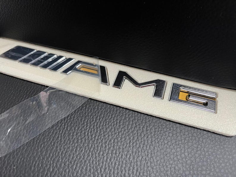 May include: Chrome AMG emblem on a white rectangular base, partially covered by a clear protective film. The emblem features the letters "AMG" in a stylized font, with a gold accent on the "G". The background is a textured black surface.