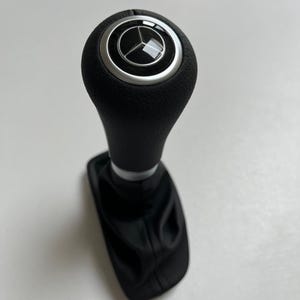 Könnte beinhalten: Schwarzer Schalthebelknauf mit silberner Zierleiste und Mercedes-Benz-Logo. Der Schalthebel ist aus schwarzem Leder gefertigt und für ein Auto konzipiert. Der Schalthebel ist ein Ersatzteil.
