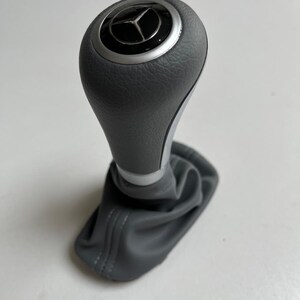 May include: Gray gear shift knob with a silver accent ring and a Mercedes-Benz logo. The gear shift is made of textured gray material and has a matching gray boot.