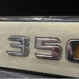 Op de afbeelding: Zilveren S350-embleem met een chromen afwerking, tegen een beige achtergrond. Het embleem is verpakt in een doorzichtige plastic verpakking. De '0' heeft een vierkant, amberkleurig midden.