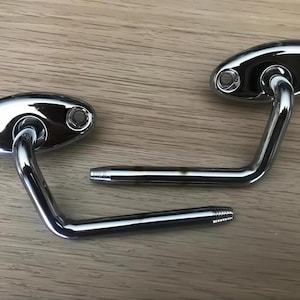 May include: Two polished chrome door or cabinet hinges. Each has an oval base with two screw holes and a curved arm ending in a threaded post. The hinges are designed for mounting on a wooden surface.