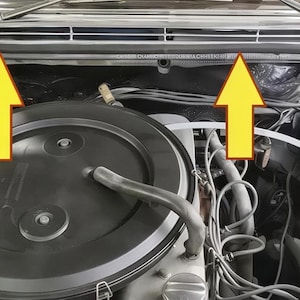 Op de afbeelding: Motorcompartiment van een oldtimer, met diverse mechanische componenten. De afbeelding toont een grote, ronde luchtfilterbehuizing, samen met bedrading, slangen en andere motoronderdelen. Het kleurenschema is een mix van zwart, zilver en diverse grijstinten.
