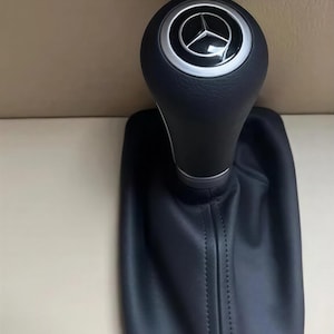 May include: Black leather gear shift knob with a silver accent ring and a Mercedes-Benz logo. The gear shift is attached to a black leather boot with stitching.