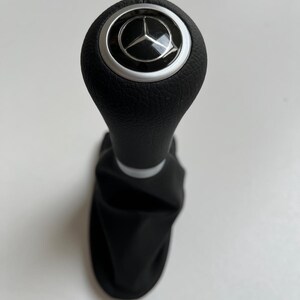May include: Black gear shift knob with a silver trim and a Mercedes-Benz logo. The knob has a textured surface and is attached to a black fabric boot.