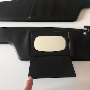 May include: Two black car sun visors. The lower visor features a rectangular mirror and a small, hinged panel. The top visor is a solid, rectangular shape. Both visors are made of a smooth, matte material.