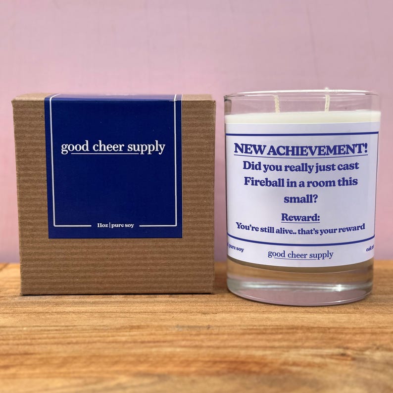 Op de afbeelding: Een sojawas kaars in een heldere glazen pot met een wit etiket met de tekst "NEW ACHIEVEMENT!". Daarnaast een bruine kartonnen doos met een blauw etiket met de tekst "good cheer supply".