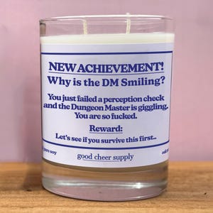 May include: A clear glass candle with a white label featuring blue text that reads "NEW ACHIEVEMENT! Why is the DM Smiling?" and other text. The candle is filled with a white wax and has two wicks. The words "pure soy" and "good cheer supply" are also visible.