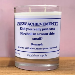 May include: A clear glass candle with a white label and blue text that reads "NEW ACHIEVEMENT! Did you really just cast Fireball in a room this small? Reward: You're still alive.. that's your reward." The candle is pure soy.