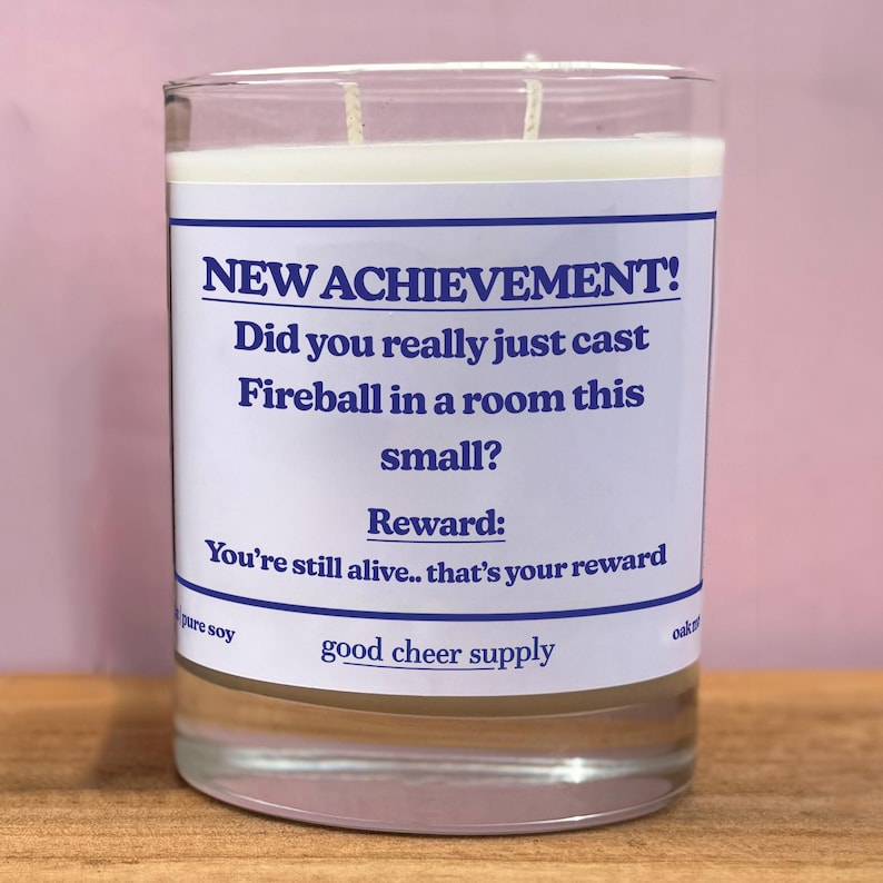 Op de afbeelding: Een heldere glazen kaars met een wit etiket en blauwe tekst. De tekst luidt: "NEW ACHIEVEMENT! Heb je net Fireball in een kamer zo klein gecast? Beloning: Je leeft nog.. dat is je beloning." De kaars is pure soja.