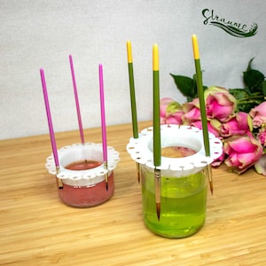 Puede incluir: Dos soportes para pinceles de plástico blanco con pinceles verdes y rosas. Los soportes están llenos de agua y tienen un borde de encaje blanco alrededor de la parte superior.