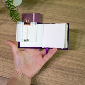 Könnte beinhalten: Ein kleines violettes Notizbuch mit einem weißen Papiereinsatz und einem goldenen Büroklammer. Das Notizbuch hat ein violettes Satinband als Lesezeichen.