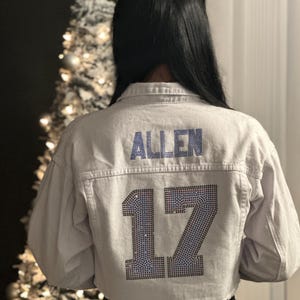 Könnte beinhalten: Weiße Jeansjacke mit einem blauen Strass-Design, auf dem hinten "ALLEN 17" steht.
