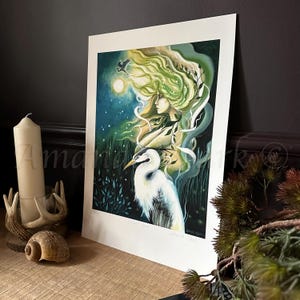 May include: A green and blue illustration of a woman with long flowing hair and a white heron. The woman is surrounded by green foliage and the moon is visible in the background.