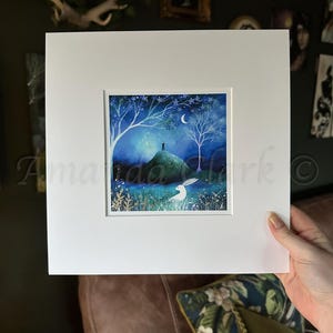 Peut inclure: Une peinture à l'aquarelle encadrée d'une colline verte avec un lapin blanc au premier plan. La colline est surmontée d'un petit bâtiment et le ciel est bleu foncé avec des étoiles et une lune croissante.