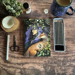 May include: A journal with a fox and woman illustration, a blue floral mug, pencils, and a small bowl on a wooden surface. The journal cover features a fox with a glowing heart and a woman with purple hair.