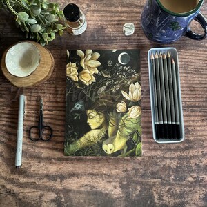 May include: A dark green journal with a colorful illustration of a woman with flowers in her hair and a crescent moon in the background. The journal is on a wooden table with other items.