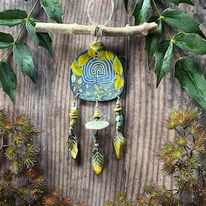 May include: A handmade wall hanging featuring a blue and yellow painted circular design with a labyrinth pattern. The artwork is adorned with leaf and bead accents, suspended from a natural wooden branch with twine.