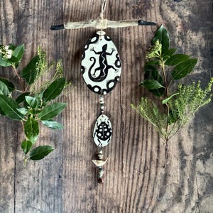 May include: A black and white wooden mobile with a lizard, ladybug, and bird design. The mobile is hanging from a piece of driftwood.