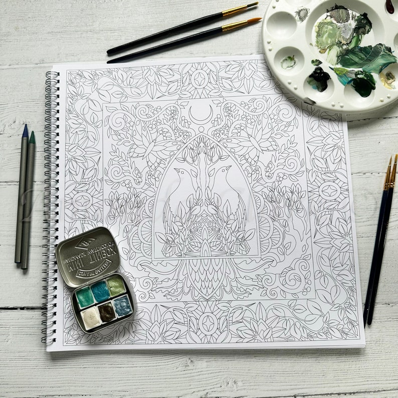 Puede incluir: Una p&aacute;gina de libro para colorear en blanco y negro con un dise&ntilde;o intrincado que presenta dos p&aacute;jaros, una luna creciente y un patr&oacute;n floral. La p&aacute;gina est&aacute; abierta sobre una mesa blanca con una lata de pinturas de acuarela y pinceles.