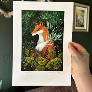 May include: A print of a fox in a forest setting. The fox is orange and white with green leaves and white flowers surrounding it. The print is signed "Amanda Clark" and titled "Manifest".