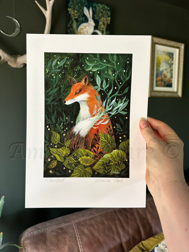 May include: A print of a fox in a forest setting. The fox is orange and white with green leaves and white flowers surrounding it. The print is signed "Amanda Clark" and titled "Manifest".