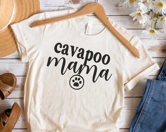 May include: Cream-colored t-shirt with the text "cavapoo mama" in black script and a paw print graphic. The shirt is displayed with blue jeans, brown sandals, and a straw hat.