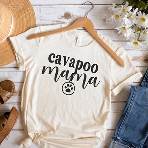 Op de afbeelding: Crèmekleurig T-shirt met de zwarte tekst "cavapoo mama" en een pootafdruk. Het shirt wordt getoond met een blauwe jeans, bruine sandalen en een strohoed.