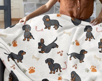 May include: A white fleece blanket with a repeating pattern of black and tan dachshund dogs, bones, and paw prints. A person is holding the blanket up, showing the design.