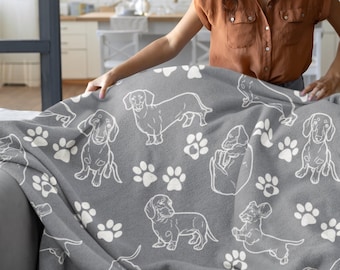 Puede incluir: Una mujer sostiene una manta gris con un patrón de dachshund blanco y huellas de patas.