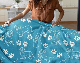 Puede incluir: Una manta de forro polar azul con un patrón de dachshund blanco y huellas de patas. Una persona está sosteniendo la manta para mostrar el diseño.