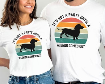 May include: A white t-shirt with a retro-style graphic of a dachshund silhouette in front of a sunset. The text "It's not a party until a wiener comes out" is printed on the shirt.
