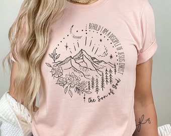 May include: A pink t-shirt with a black graphic design of a mountain range, flowers, and stars. The text "BEHOLD I AM A DISCIPLE OF JESUS CHRIST, the Son of God" is printed on the shirt.