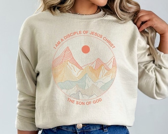 May include: A beige sweatshirt with a graphic of mountains and a sun. The graphic is surrounded by a circle with the text "I am a disciple of Jesus Christ, the son of God."