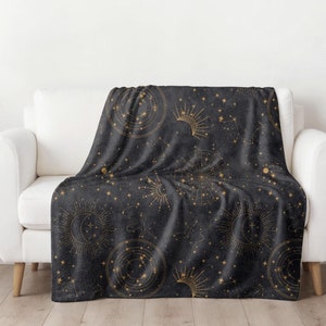 May include: A dark gray throw blanket with a gold celestial pattern featuring stars, moons, and constellations. The blanket is draped over a white couch.