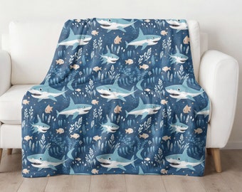 May include: A blue throw blanket with a repeating pattern of white sharks, orange fish, and blue seaweed. The blanket is draped over a white couch.