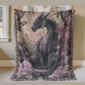 May include: A woven throw blanket featuring a detailed illustration of a black dragon amidst a forest of purple and pink flowers and foliage. The dragon has a detailed, scaled body and is surrounded by trees and flowers. The blanket has a fringed edge.