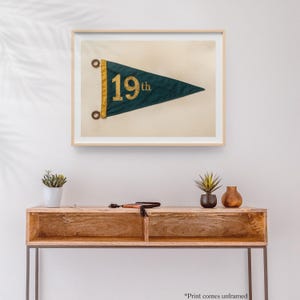 May include: A framed print of a vintage green pennant with gold lettering that reads "19th". The pennant is displayed on a wooden console table with two small potted plants.