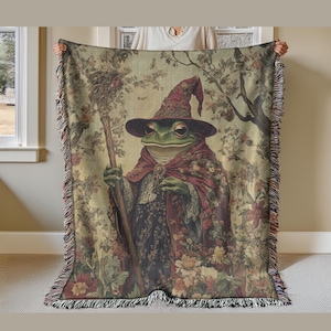Puede incluir: Una manta de tiro de tapicería con una rana verde que lleva un sombrero de mago rojo y morado y sostiene un bastón. La rana está rodeada de flores y follaje en un estilo vintage.