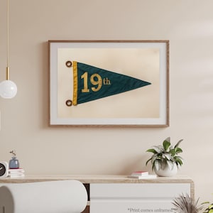May include: A framed print of a vintage green pennant with gold lettering that reads "19th". The pennant is hanging on a light beige wall.