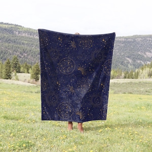 May include: A navy blue throw blanket with a gold celestial pattern featuring stars, moons, and suns. The blanket is being held up in front of a green grassy field with mountains in the background.
