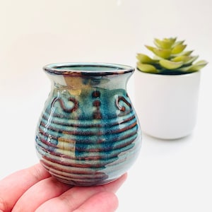 May include: A small, bulbous ceramic vase featuring a glossy glaze in shades of deep teal, blue, and earthy brown. The surface is decorated with horizontal ridges and subtle swirling patterns, finished with a smooth, flared rim at the top.