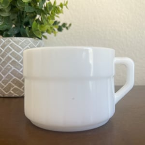 Puede incluir: Una taza de vidrio opalino blanco con una sección superior lisa y una sección inferior con nervaduras verticales. La taza tiene un asa angular y resistente unida al lateral, ofreciendo un diseño limpio y sencillo para bebidas.