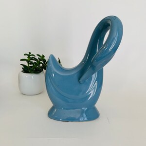 May include: A glossy, dusty blue ceramic swan-shaped planter or decorative vessel with a smooth, curved neck and a wide, stable base. The piece features a hollow center and a reflective finish, showcasing a solid, monochromatic color throughout.